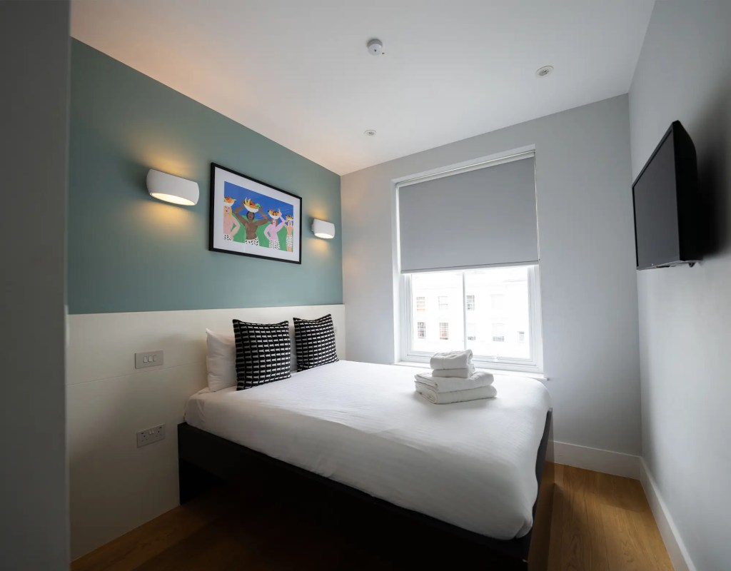 A stylish one-bedroom serviced apartment overlooking Portobello Road, Notting Hill, with a staircase leading to the upper level, a modern sleeping area with a plush double bed, wooden floors, underfloor heating, and an entrance to an elegant private bathroom with a shower