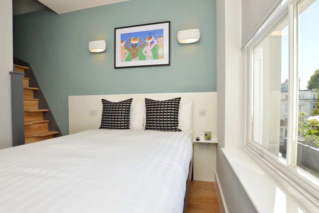 A stylish one-bedroom serviced apartment overlooking Portobello Road, Notting Hill, with a staircase leading to the upper level, a modern sleeping area with a plush double bed, wooden floors, underfloor heating, and an entrance to an elegant private bathroom with a shower