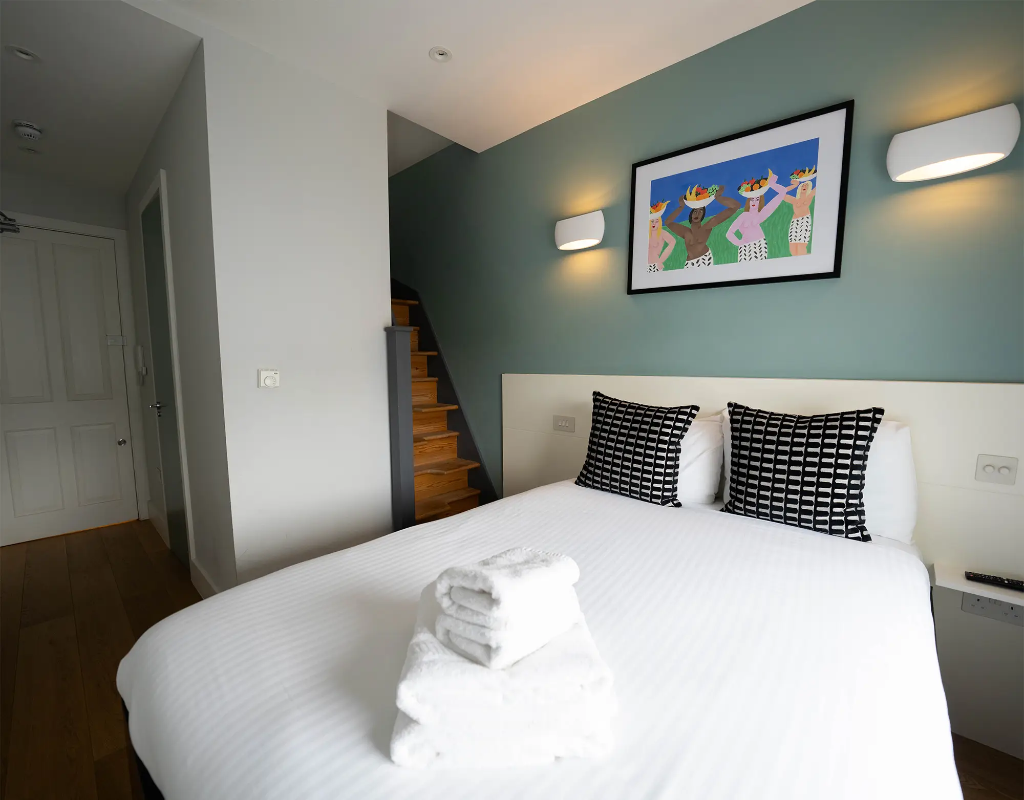 A stylish one-bedroom serviced apartment overlooking Portobello Road, Notting Hill, with a staircase leading to the upper level, a modern sleeping area with a plush double bed, wooden floors, underfloor heating, and an entrance to an elegant private bathroom with a shower
