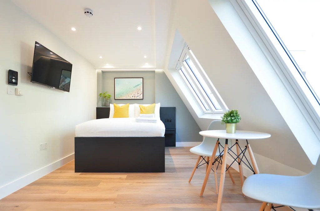 Sleeping area with cozy double bed and light-filled roof light windows. Hardwood floor, dining area with table and two chairs, and TV entertainment unit in a Marylebone standard studio apartment at 162 New Cavendish St, London