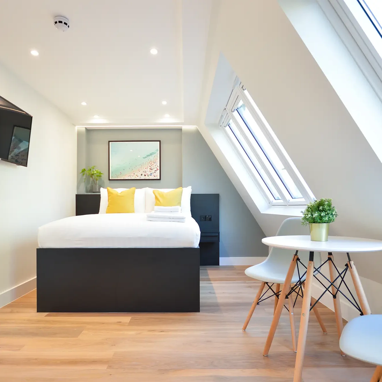 Sleeping area with cozy double bed and light-filled roof light windows. Hardwood floor, dining area with table and two chairs, and TV entertainment unit in a Marylebone standard studio apartment at 162 New Cavendish St, London